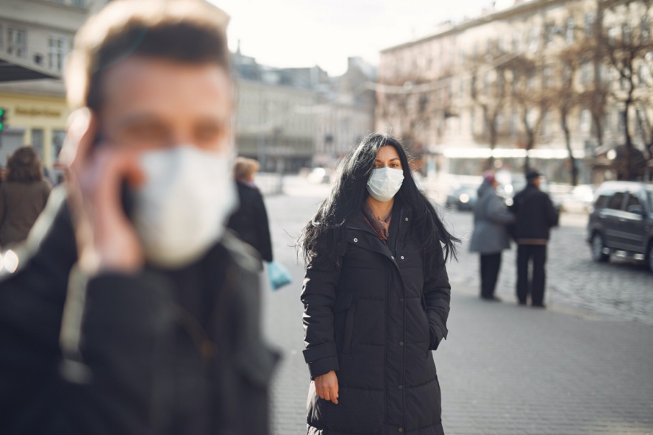 People in the street wearing masks. Man in foreground is blurry, focal point is woman walking with mask on and buildings in the background. 